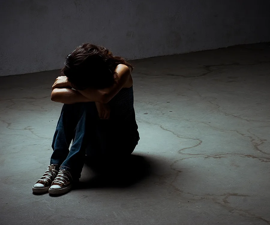 A person sitting on the floor in a dimly lit room, hugging their knees, with a sense of solitude and distress.