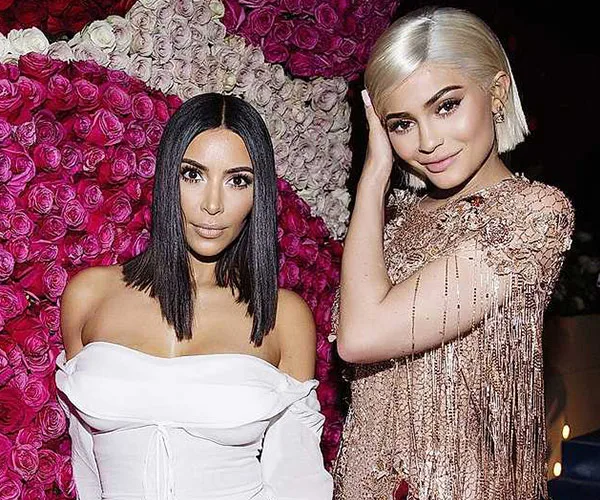 Two women posing together in front of a wall of pink roses, one in a white outfit, the other in a shimmering dress.