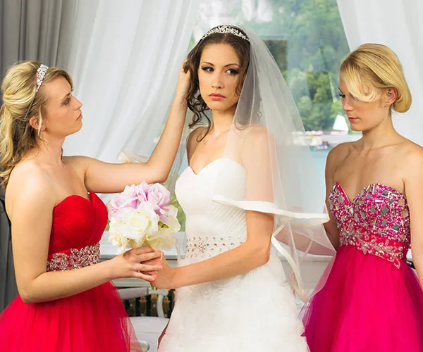 Bridal party scene with a focused bride in a white gown and two bridesmaids in red dresses adjusting her veil and holding flowers.