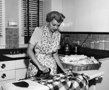 Woman ironing a plaid shirt in a vintage kitchen, wearing an apron, with a basket of laundry nearby.