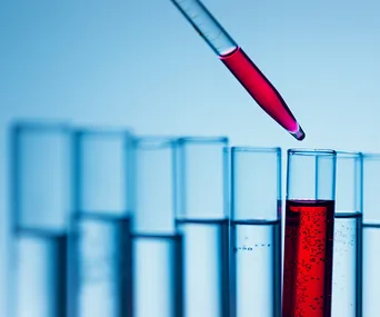 Pipette drips red liquid into a test tube among several others in a lab setting, against a blue gradient background.