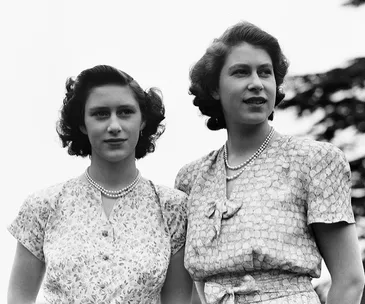 Two women standing outdoors in vintage dresses, wearing pearl necklaces, looking into the distance.