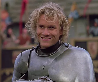 A knight in armor smiles warmly, with curly hair, set against a medieval background with spectators.