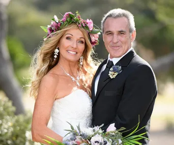 A couple in wedding attire stands outdoors; the woman wears a floral crown, and the man is in a suit with a floral boutonniere.