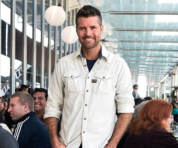 Man in a white shirt smiling in a busy restaurant under a glass ceiling with hanging paper lanterns.