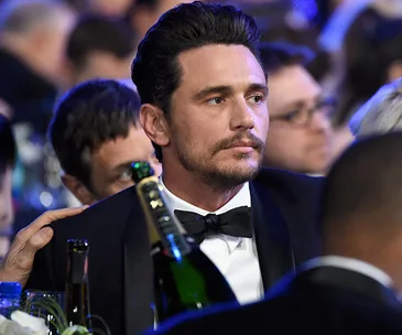 A man in a tuxedo sits at a formal event, surrounded by people and champagne bottles.