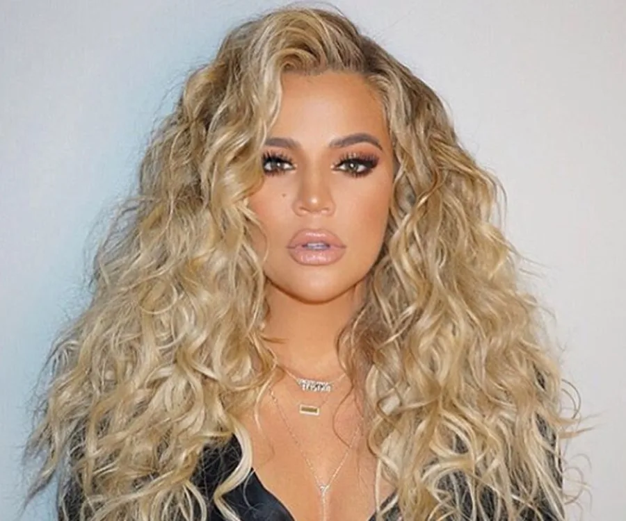 Blonde woman with long, curly hair and makeup, wearing a necklace and a black top, posing against a neutral background.
