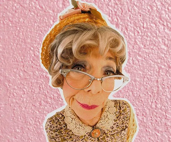 Elderly woman with glasses and a warm hat, smiling slightly, wearing vintage clothing against a textured pink background.