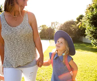 Mother and daughter holding hands, the child wearing a blue sun hat and backpack, smiling outdoors.