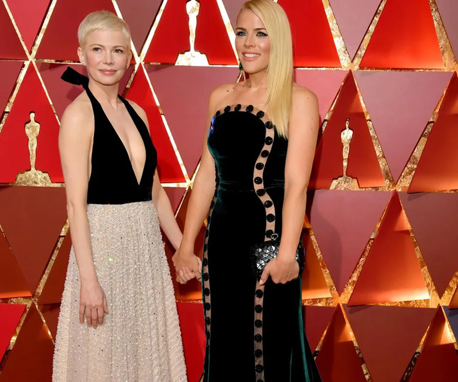 Two women holding hands at a formal event in front of a red and gold geometric backdrop.