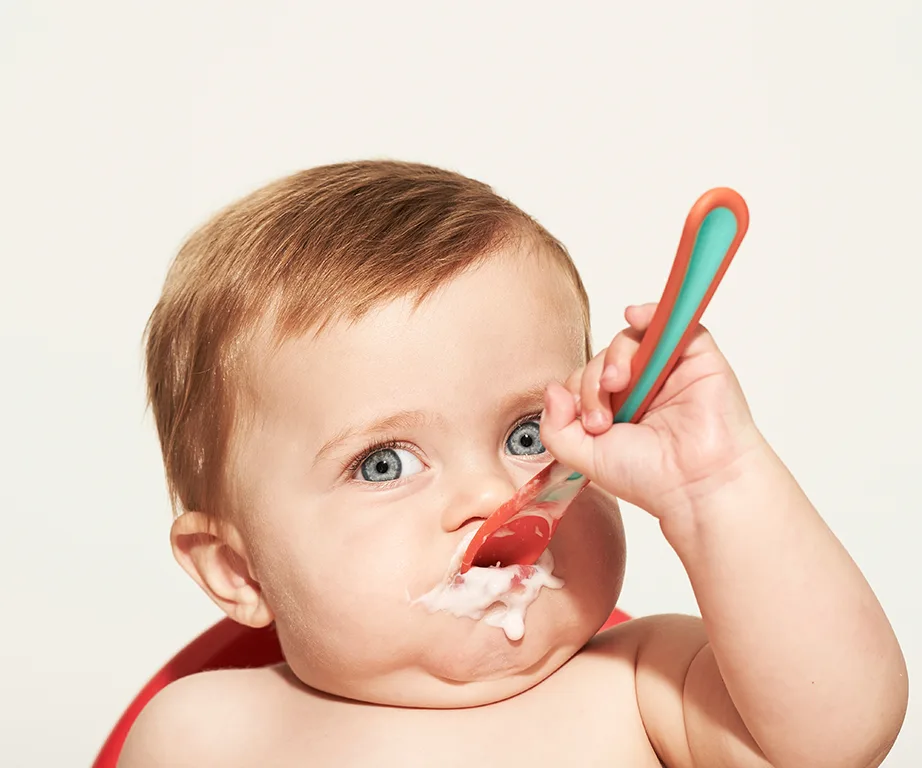 Baby eating food with a spoon, some food on face, bright eyes and light brown hair, against a neutral background.