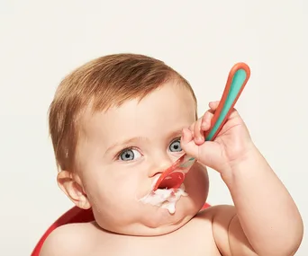 Baby eating food with a spoon, some food on face, bright eyes and light brown hair, against a neutral background.