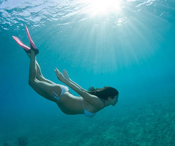 Pregnant woman snorkeling underwater with sunlight streaming down, wearing a white bikini and pink fins.