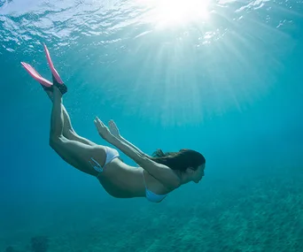 Pregnant woman snorkeling underwater with sunlight streaming down, wearing a white bikini and pink fins.