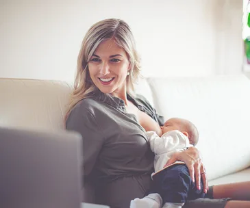 Woman multitasking; breastfeeding baby while working on a laptop, smiling and seated on a couch.