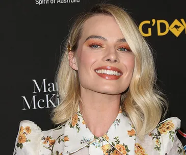 Blonde woman in floral dress smiling on the G'Day USA red carpet event backdrop.