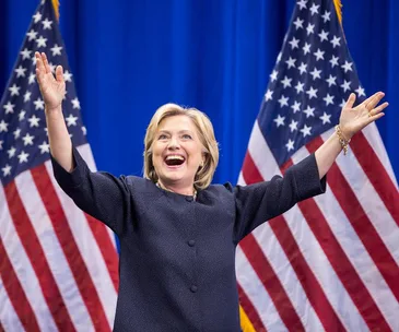 Hillary Clinton smiling with arms raised, standing in front of two American flags on a blue background.