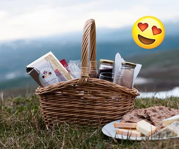 Picnic basket with snacks, jars, and chocolate on grass; heart-eyes emoji in the sky, mountains in the background.
