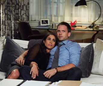 A couple sits closely on a sofa, appearing thoughtful and relaxed, with a home office in the background.