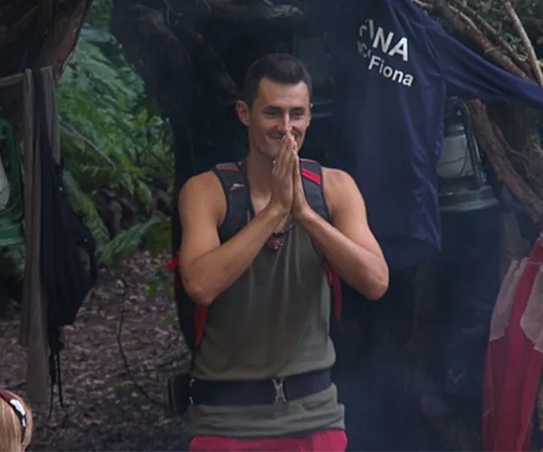 Individual in camp gear standing with hands together, surrounded by nature, with clothing hanging in background.