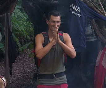 Individual in camp gear standing with hands together, surrounded by nature, with clothing hanging in background.
