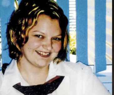 Portrait of a smiling woman with short, curly hair and a white blouse in front of a window with vertical blinds.