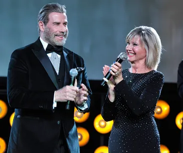 John Travolta and Olivia Newton-John on stage, holding microphones and smiling at each other.