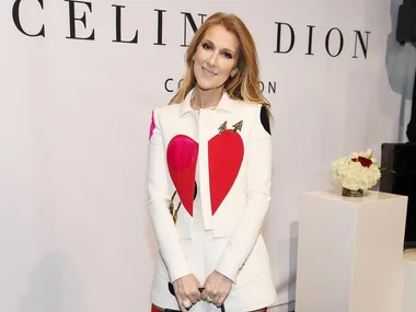 Celine Dion at a promotional event, wearing a white jacket with heart designs, in front of a branded backdrop.