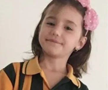 Young girl smiling, wearing a school uniform with a pink bow in her hair.