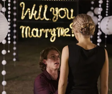 A man on one knee proposes to a woman, with "Will you marry me?" in lights behind them.