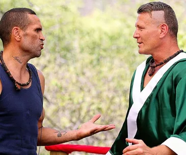 Two men are having a conversation outdoors; one in a tank top gesturing, the other in a green robe listening.