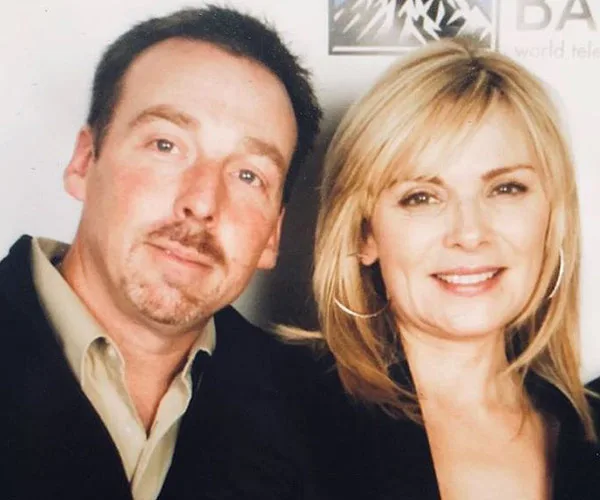 Two people posing together, with one man in a suit and a woman with blonde hair smiling at the camera.