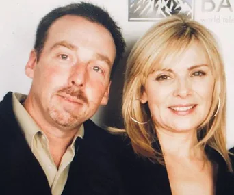 Two people posing together, with one man in a suit and a woman with blonde hair smiling at the camera.