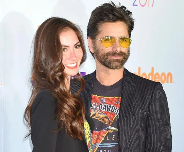 A couple smiling at an event, with the man wearing sunglasses and a graphic T-shirt under a blazer.