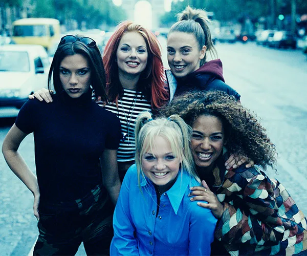 Five women posing on a street, all smiling, with one wearing red and black stripes and one in a blue shirt, looking cheerful.