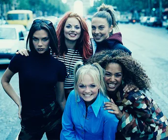 Five women posing on a street, all smiling, with one wearing red and black stripes and one in a blue shirt, looking cheerful.