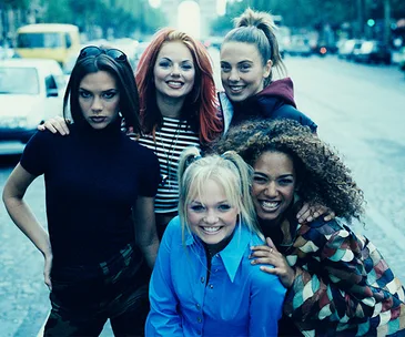 Five women posing on a street, all smiling, with one wearing red and black stripes and one in a blue shirt, looking cheerful.