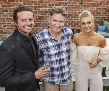 Three people standing together, smiling and holding drinks in an outdoor setting with a brick wall backdrop.