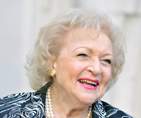 Elderly woman with white hair, wearing a patterned jacket and pearl necklace, smiling brightly outdoors.