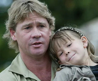 Father holding young daughter close, both smiling softly outdoors; daughter wears a headband.