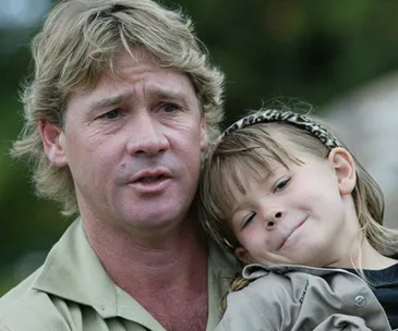 Father holding young daughter close, both smiling softly outdoors; daughter wears a headband.