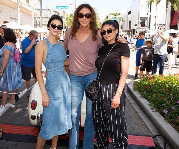 Three people posing outdoors, two in sunglasses, with a car and crowd in the background.