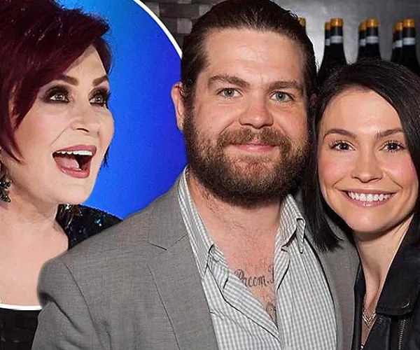 Sharon Osbourne smiling alongside a man in a suit and a woman with black hair, posing for a photo.