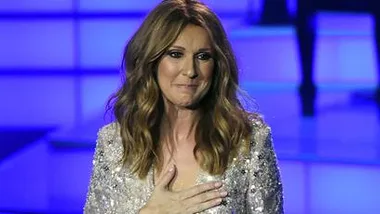 Celine Dion, visibly emotional, holds her hand to her chest while on stage, wearing a sparkling outfit.