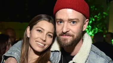 Two people smiling, one wearing a red beanie, in a close embrace at an event, with greenery in the background.