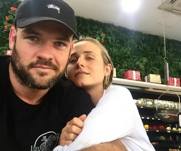 Couple smiling at the camera; man in black cap, woman leaning on him, green foliage and gifts in the background.