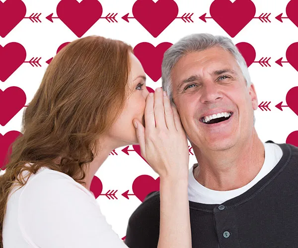 A woman whispers to a smiling man against a background of red hearts and arrows.