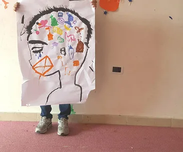 Child holding colorful drawing of a face with various symbols on a wall background, standing on a carpeted floor.