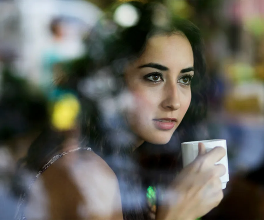 Woman sipping coffee indoors, gazing thoughtfully through a glass window with reflections.