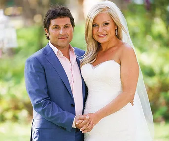 A couple standing together outdoors, the woman in a wedding dress and the man in a blue suit.